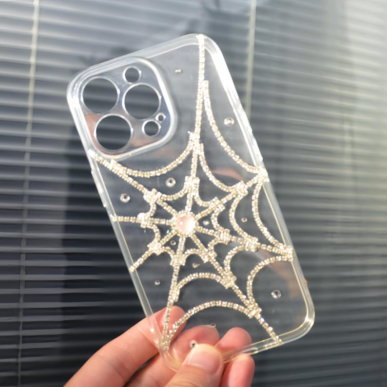 May include: A clear phone case with a sparkly spiderweb design. The web is made of rhinestones and there is a larger pink rhinestone in the center.