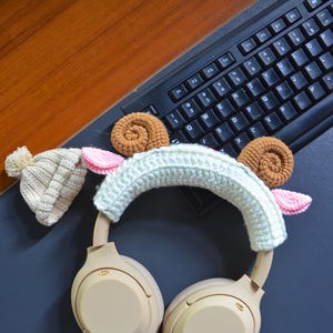 K&ouml;nnte beinhalten: Beigefarbene kabellose Kopfh&ouml;rer mit einem wei&szlig;en H&auml;kelband mit braunen und rosa Akzenten. Das Band hat ein Schafdesign mit Ohren.