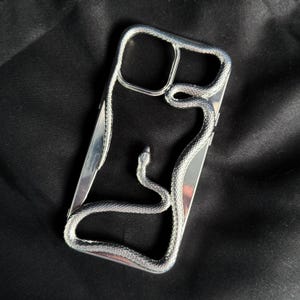 Puede incluir: Una funda de teléfono plateada con un diseño de serpiente. La serpiente está envuelta alrededor de la funda y su cabeza es visible en la parte superior.