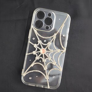 May include: Clear phone case with a spiderweb design made of rhinestones. The spiderweb has a pink rhinestone in the center.