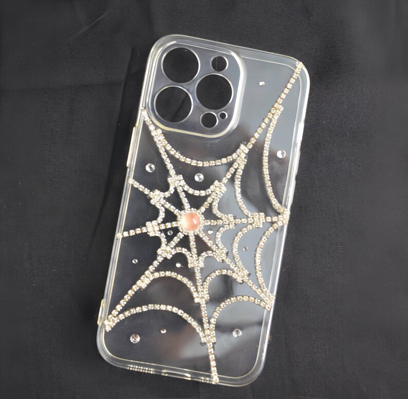 May include: Clear phone case with a spiderweb design made of rhinestones. The spiderweb has a pink rhinestone in the center.