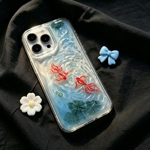 May include: A clear smartphone case featuring a textured, rippled water design with two red goldfish swimming, green lily pads, a small white flower-shaped charm, and a light blue bow-shaped charm attached to the side of the case.