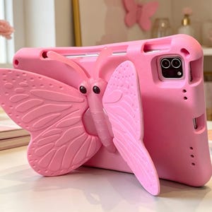 May include: A bright pink tablet case with a 3D butterfly design. The butterfly has detailed wings and a textured body. The case is designed to protect the tablet and features a camera cutout. The overall aesthetic is playful and girly.