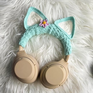 May include: Beige wireless headphones with a light blue crocheted cat ear headband with a pink flower accent.