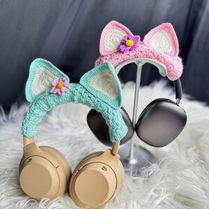 May include: Two pairs of headphones with crocheted cat ear covers. One pair is pink with a purple flower and the other is teal with a pink flower.