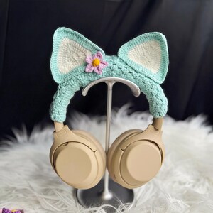 May include: A pair of beige headphones with a crocheted headband featuring a light blue and white cat ear design with a pink and yellow flower accent.