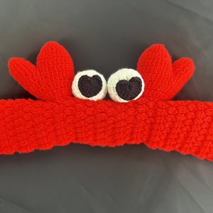 Op de afbeelding: Rode gebreide hoofdband met twee witte ogen en zwarte pupillen, in de vorm van een krab.
