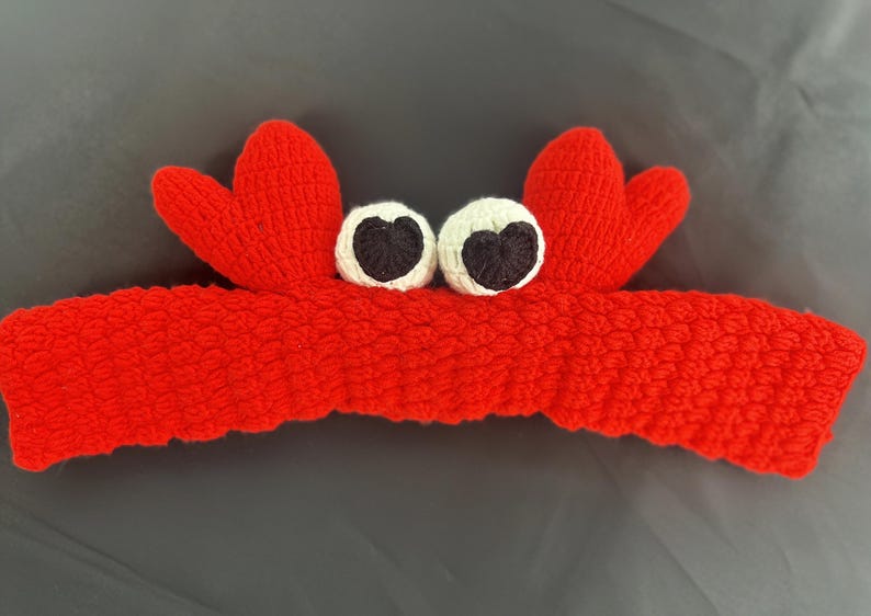 Op de afbeelding: Rode gebreide hoofdband met twee witte ogen en zwarte pupillen, in de vorm van een krab.