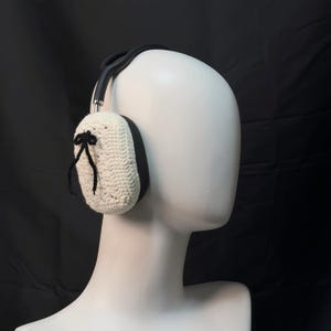 May include: White crocheted earmuffs with black ribbon accents and a black headband. The earmuffs are worn on a white mannequin head.