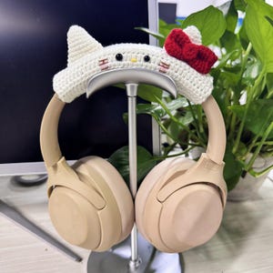 May include: A white crocheted headphone stand with a red bow and black eyes. The stand is holding a pair of beige headphones.