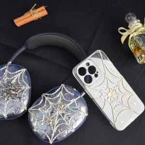May include: A pair of headphones and a phone case decorated with a spiderweb design and rhinestones. The headphones and phone case are both clear and have a silver spiderweb design. The rhinestones are scattered throughout the spiderweb design.