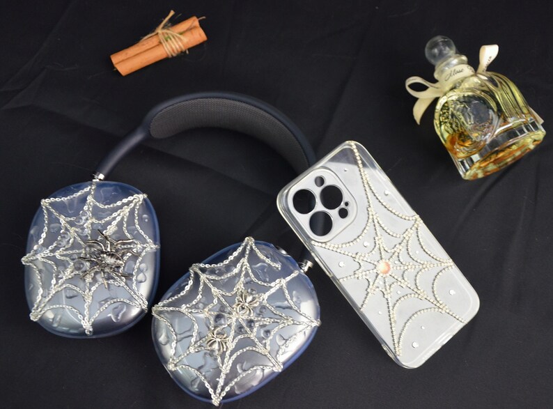 May include: A pair of headphones and a phone case decorated with a spiderweb design and rhinestones. The headphones and phone case are both clear and have a silver spiderweb design. The rhinestones are scattered throughout the spiderweb design.