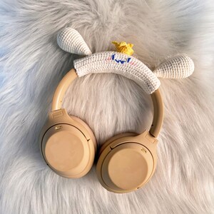 May include: Tan wireless headphones with a white crocheted headband featuring a cartoon face with blue and yellow accents. The headband has two white crocheted ears and a yellow crown. The headphones rest on a fluffy white surface.