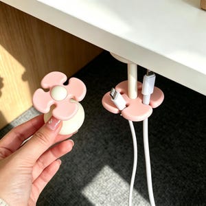 May include: A light pink and white flower-shaped cable organizer. The organizer is designed to hold multiple charging cables. One is held by a hand. Another is attached to a desk. The cables are white.