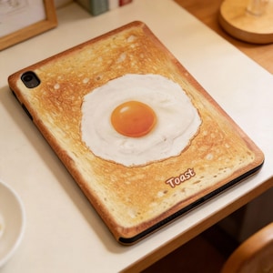 May include: A tablet case with a printed design of a slice of toast topped with a fried egg. The egg has a bright orange yolk and white egg whites. The word "Toast" is printed in a brown font on the bottom right corner.