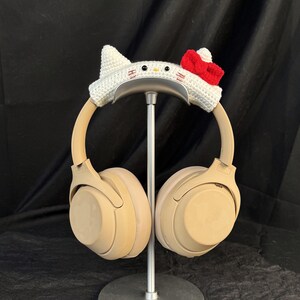 May include: Beige over-ear headphones with a white crocheted Hello Kitty headband. The headband has a red bow.