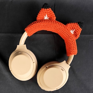 May include: Beige wireless headphones with a crocheted fox ear headband in orange and black.