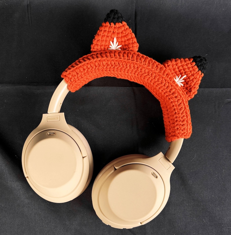 May include: Beige wireless headphones with a crocheted fox ear headband in orange and black.