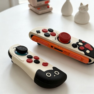 Könnte beinhalten: Zwei weiße und orangefarbene Gamecontroller mit schwarzen und roten Knöpfen. Ein Controller hat ein Katzen-Gesichts-Design am Ende. Die Controller sind für eine Spielkonsole konzipiert. Die Controller liegen auf einer weißen Oberfläche.