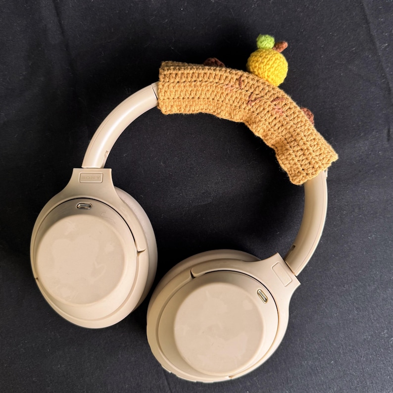 May include: Beige wireless headphones with a crocheted brown and yellow headband with a small yellow and green crocheted apple on top.