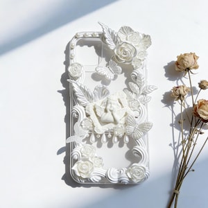 Könnte beinhalten: Transparente Handyhülle mit weißen Blumen- und Schmetterlingsverzierungen. Mit einer geformten Engelsfigur, Rosen und perlenartigen Akzenten. Die Hülle ist in einem dekorativen, verzierten Stil gestaltet.