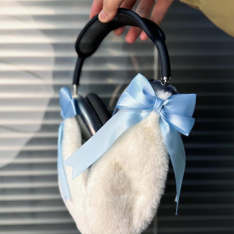 May include: A pair of headphones with a white faux fur earmuff cover and a light blue satin bow. The headphones are black and have a silver accent.
