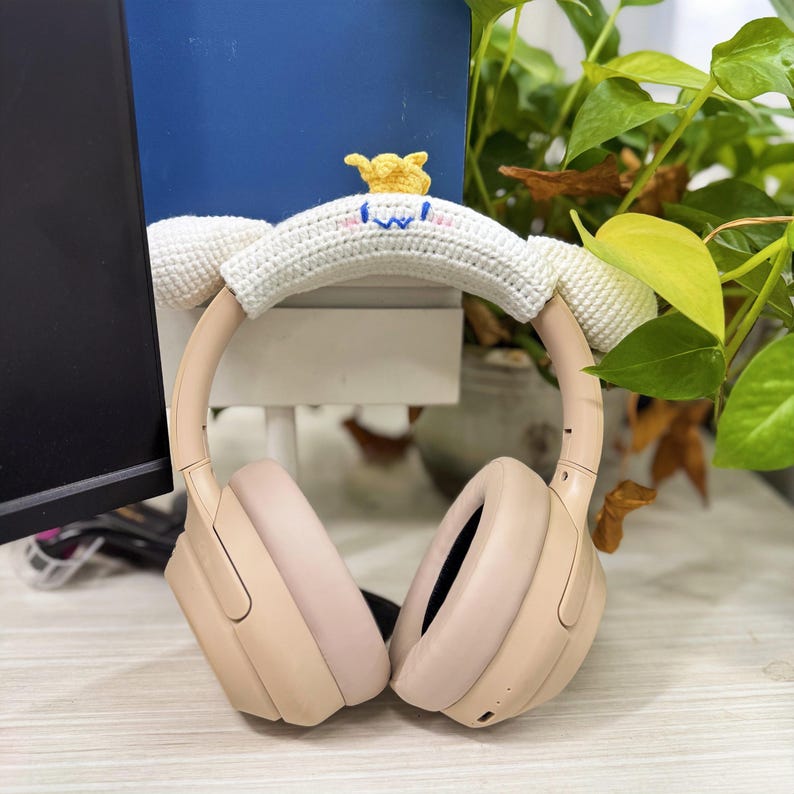 May include: Blush-colored over-ear headphones with a white crocheted headband cover featuring a cartoon face and a yellow crown. The headphones are resting on a light-colored surface, with a blue background and greenery.