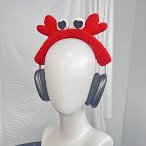 Op de afbeelding: Een rood gebreid krabbenhoofdband met witte ogen en zwarte pupillen. De hoofdband wordt gedragen op een wit mannequinhoofd met zwarte koptelefoon.