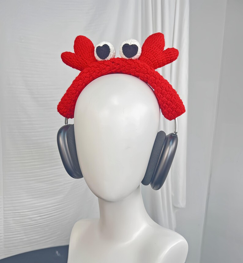 Op de afbeelding: Een rood gebreid krabbenhoofdband met witte ogen en zwarte pupillen. De hoofdband wordt gedragen op een wit mannequinhoofd met zwarte koptelefoon.
