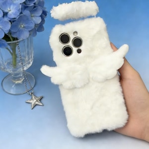 Könnte beinhalten: Weißes, flauschiges Handycase mit Engelsflügeln und Heiligenschein. Das Case hat ein weiches, strukturiertes Aussehen und einen Kameraausschnitt. Ein sternförmiger Anhänger hängt in der Nähe einer Vase mit blauen Blumen.