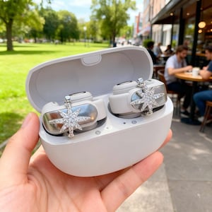 Puede incluir: Auriculares inalámbricos blancos con detalles plateados y un diseño de estrella, presentados en un estuche de carga a juego. Los auriculares están adornados con cristales brillantes, añadiendo un toque de elegancia.