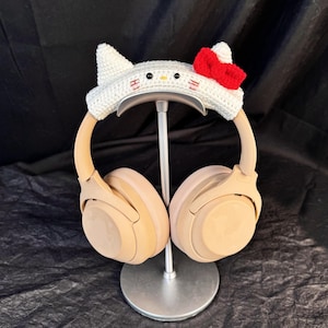 May include: Beige wireless headphones with a white crocheted headband featuring a Hello Kitty design with a red bow. The headphones are on a silver stand.