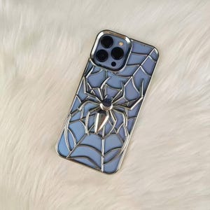 May include: A blue phone case with a silver spider web design and a silver spider in the center. The case has a silver border around the camera and the edges of the phone.