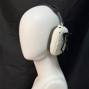 May include: White crocheted earmuffs with black ties attached to a black headband. The earmuffs are worn on a white mannequin head.