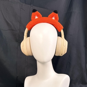 May include: Orange crocheted fox headband with black ears, worn over a pair of beige over-ear headphones.