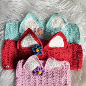 May include: Three crocheted headbands with cat ears and flower embellishments. The headbands are in shades of pink, red, and light blue.