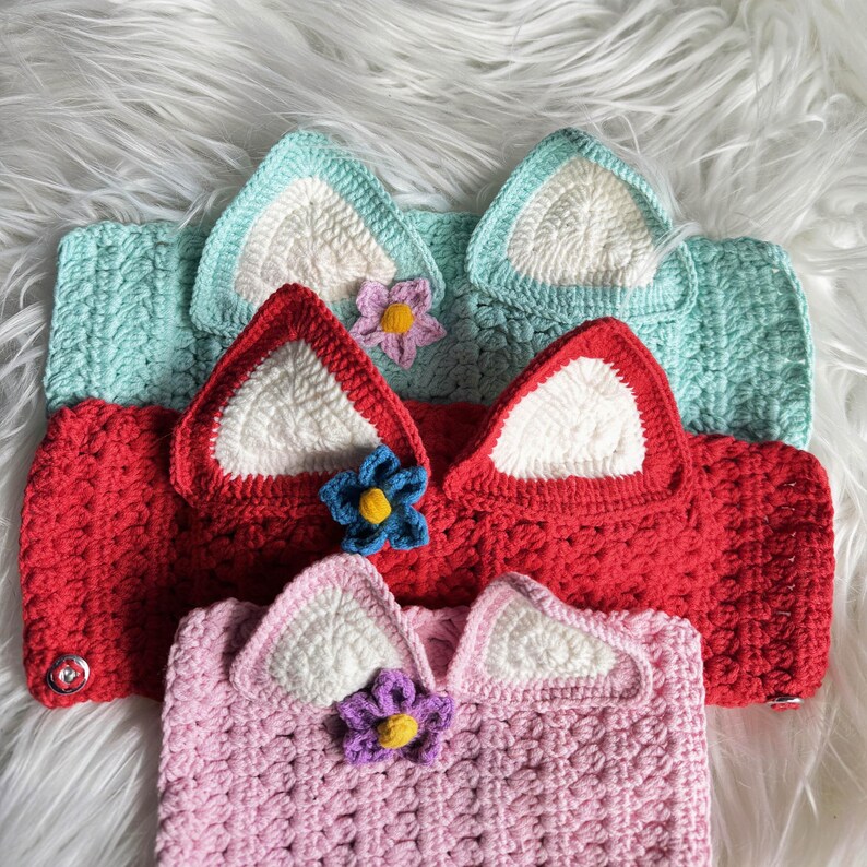 May include: Three crocheted headbands with cat ears and flower embellishments. The headbands are in shades of pink, red, and light blue.