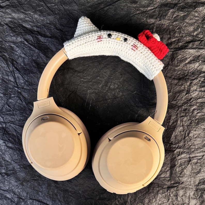 May include: Beige wireless headphones with a white crocheted cat ear headband. The headband has red accents and black embroidered eyes.