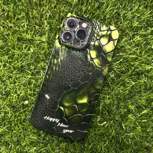 Puede incluir: Una funda de teléfono verde y negra con un patrón de escamas de reptil. La funda tiene un acabado brillante y presenta el texto "Happy New Year" en escritura blanca. La funda está sobre hierba verde.