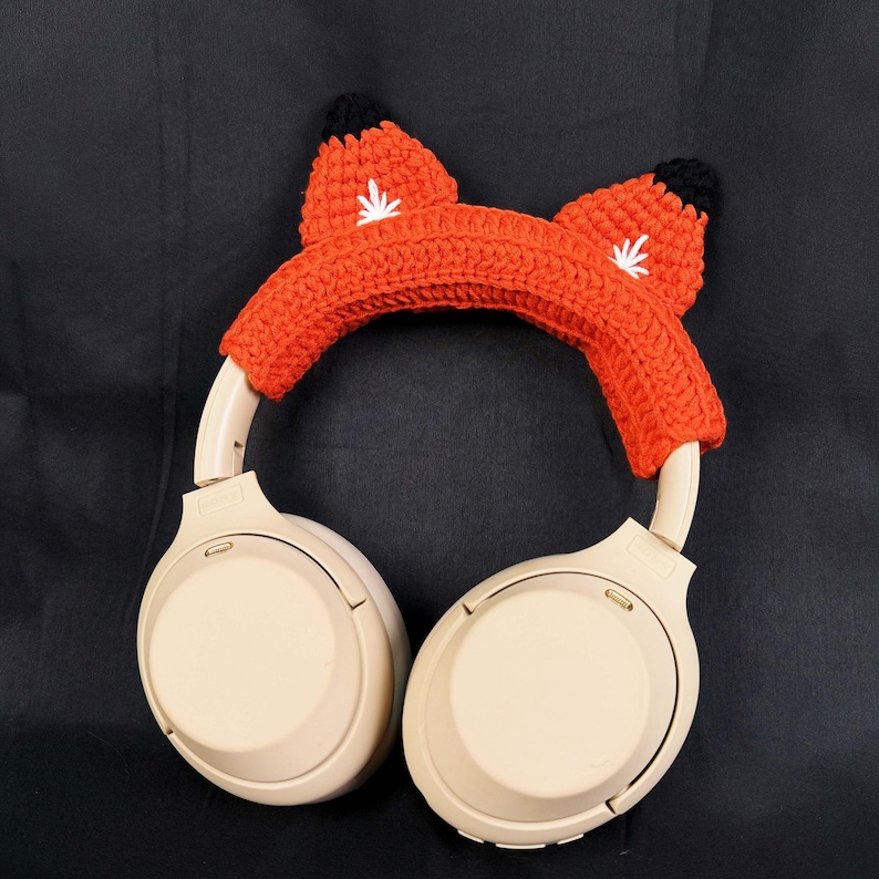 May include: Beige wireless headphones with a crocheted orange fox ear headband. The headband has black felt ears and white embroidered details.