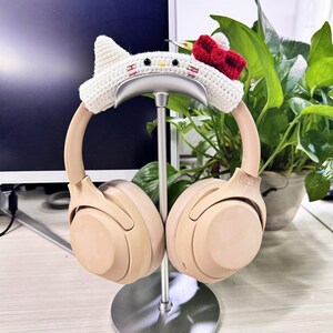 May include: Beige wireless headphones with a white crocheted headband featuring a Hello Kitty design. The headphones are on a silver stand.