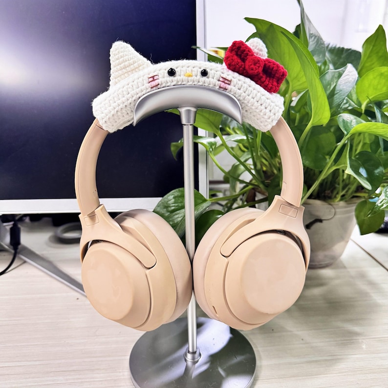 May include: Beige wireless headphones with a white crocheted headband featuring a Hello Kitty design. The headphones are on a silver stand.