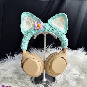 May include: A pair of beige headphones with a crocheted cat ear headband. The headband is light blue with a white flower accent.