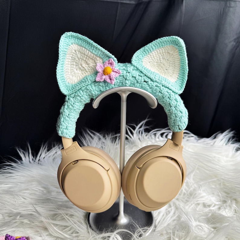 May include: A pair of beige headphones with a crocheted cat ear headband. The headband is light blue with a white flower accent.