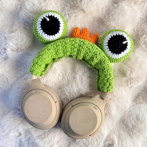 Puede incluir: Funda de auriculares de ganchillo verde en forma de rana con ojos blancos y negros y una corona naranja. La funda está en unos auriculares beige.