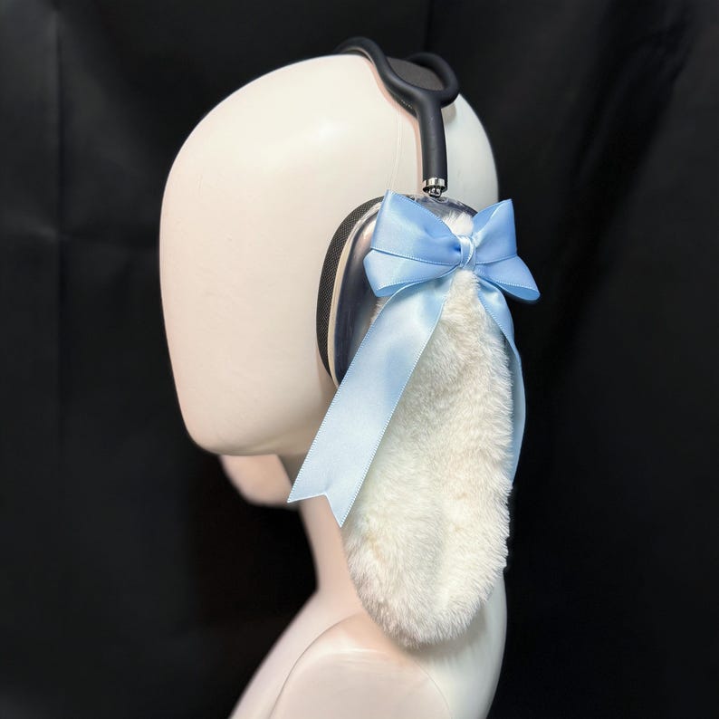 May include: A pair of white furry bunny ears with a light blue bow attached to a silver headband. The headband is worn on a white mannequin head.