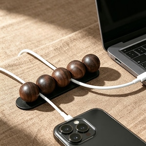 May include: A black cable organiser featuring five dark brown wooden spheres arranged in a row on a flat base. The device is positioned to hold white charging cables, with one cable connected to a smartphone and another to a laptop computer.