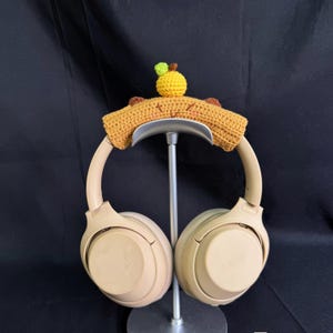 May include: Beige wireless headphones with a crocheted brown and yellow headband with a small yellow apple on top. The headphones are on a metal stand.