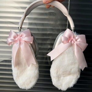 May include: White faux fur headphones with pink satin bows on each earcup. The headband is silver.