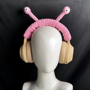 Op de afbeelding: Roze gehaakt hoofdband met twee cartoonogen, ontworpen om over koptelefoons te passen. Het hoofdband wordt gedragen door een wit mannequin.